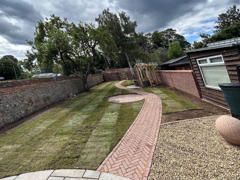 After — the same spot with new striped turf, a curving herringbone brick path, fresh gravel and an oak pergola